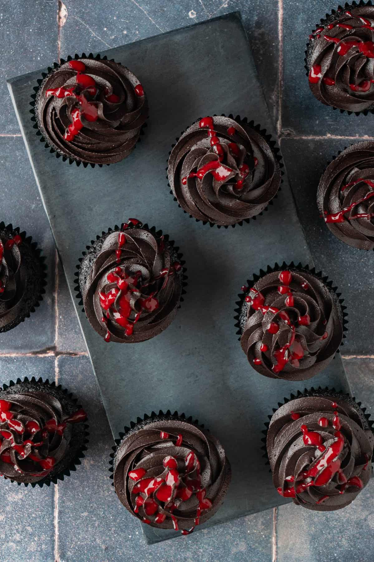 Overhead view of red and black Halloween cupcakes with swirled black frosting and raspberry drizzle on a slate board.
