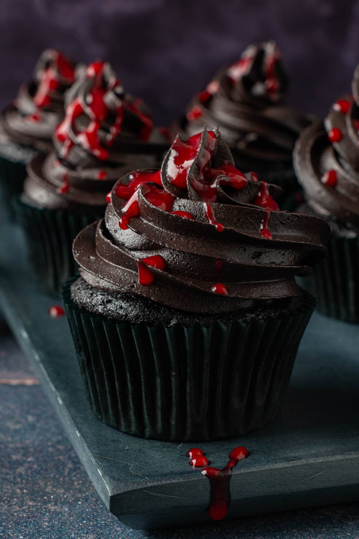 Black Halloween cupcakes with swirled black cocoa frosting and a red raspberry drizzle on a dark slate board.