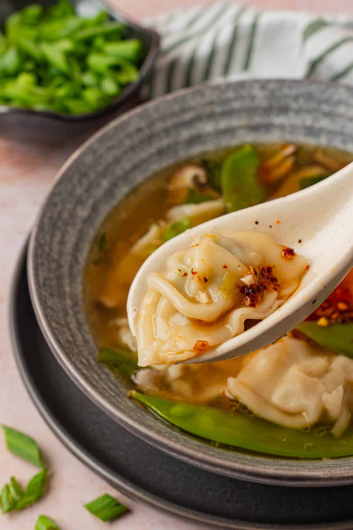 Close-up of a spoon lifting a potsticker topped with chili crisp out of a bowl of soup, with snow peas and more potstickers visible in the bowl below.