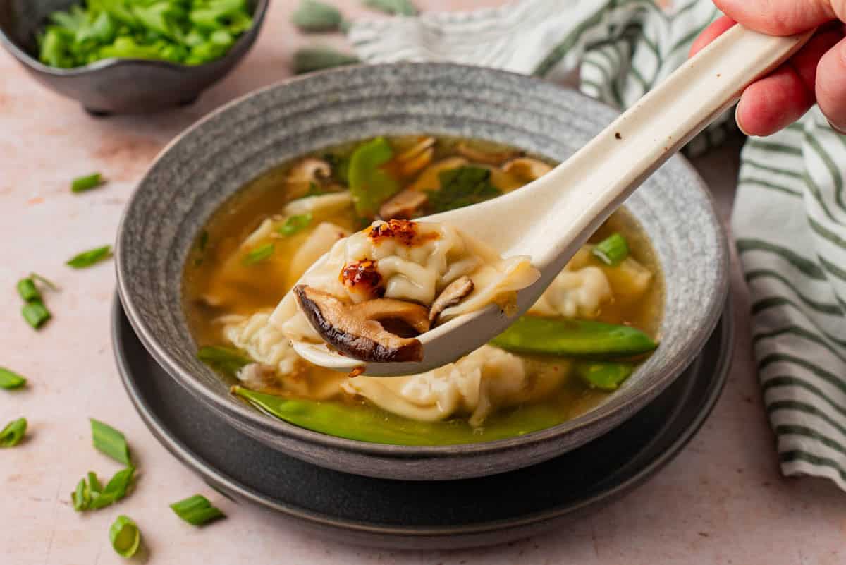 A hand holding a spoonful of potstickers, mushrooms and chili crisp over a bowl of soup, with a bowl of chopped green onions in the background.