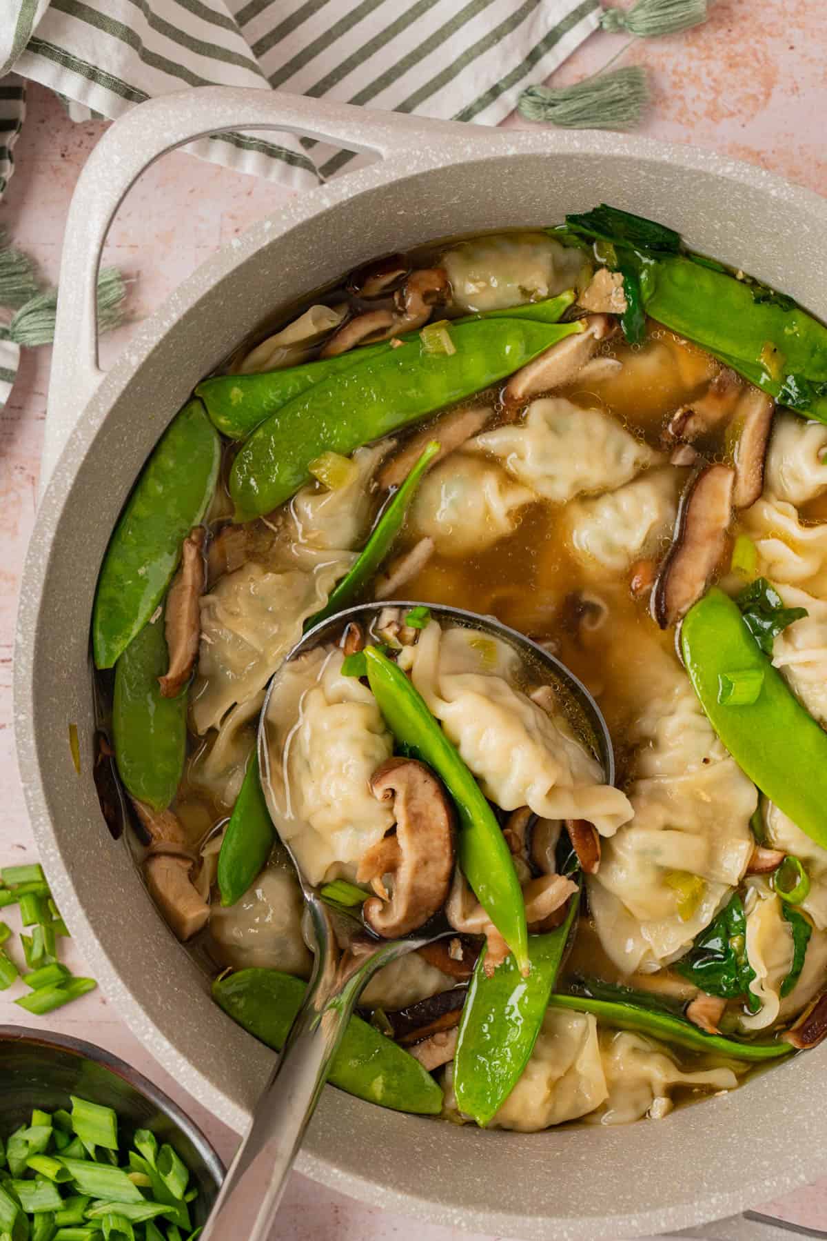 Overhead view of potsticker soup in a large gray pot, filled with mushroom-studded broth, snow peas, potstickers and sliced green onions, with a ladle scooping up a serving.