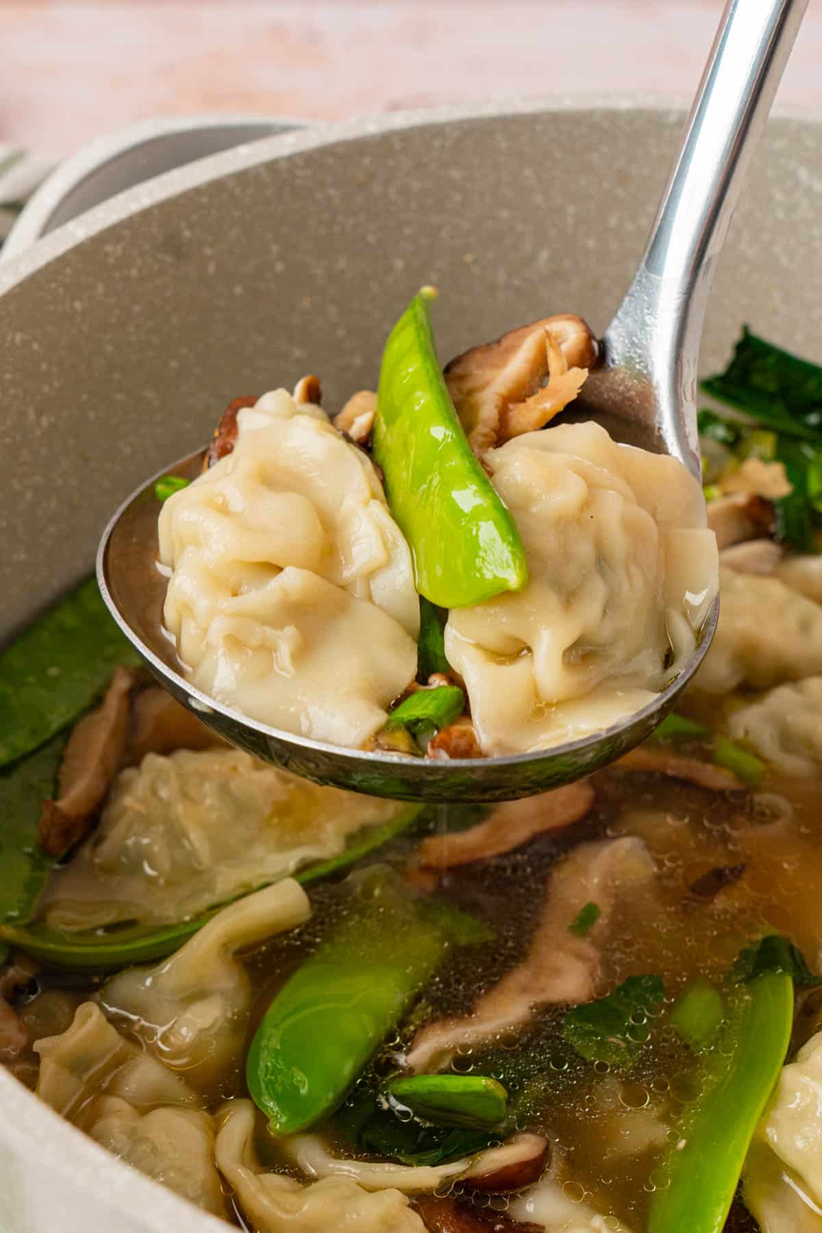 Close-up of a ladle lifting two potstickers, mushrooms and a snow pea out of the soup pot, with more potstickers, mushrooms and snow peas visible in the broth below.