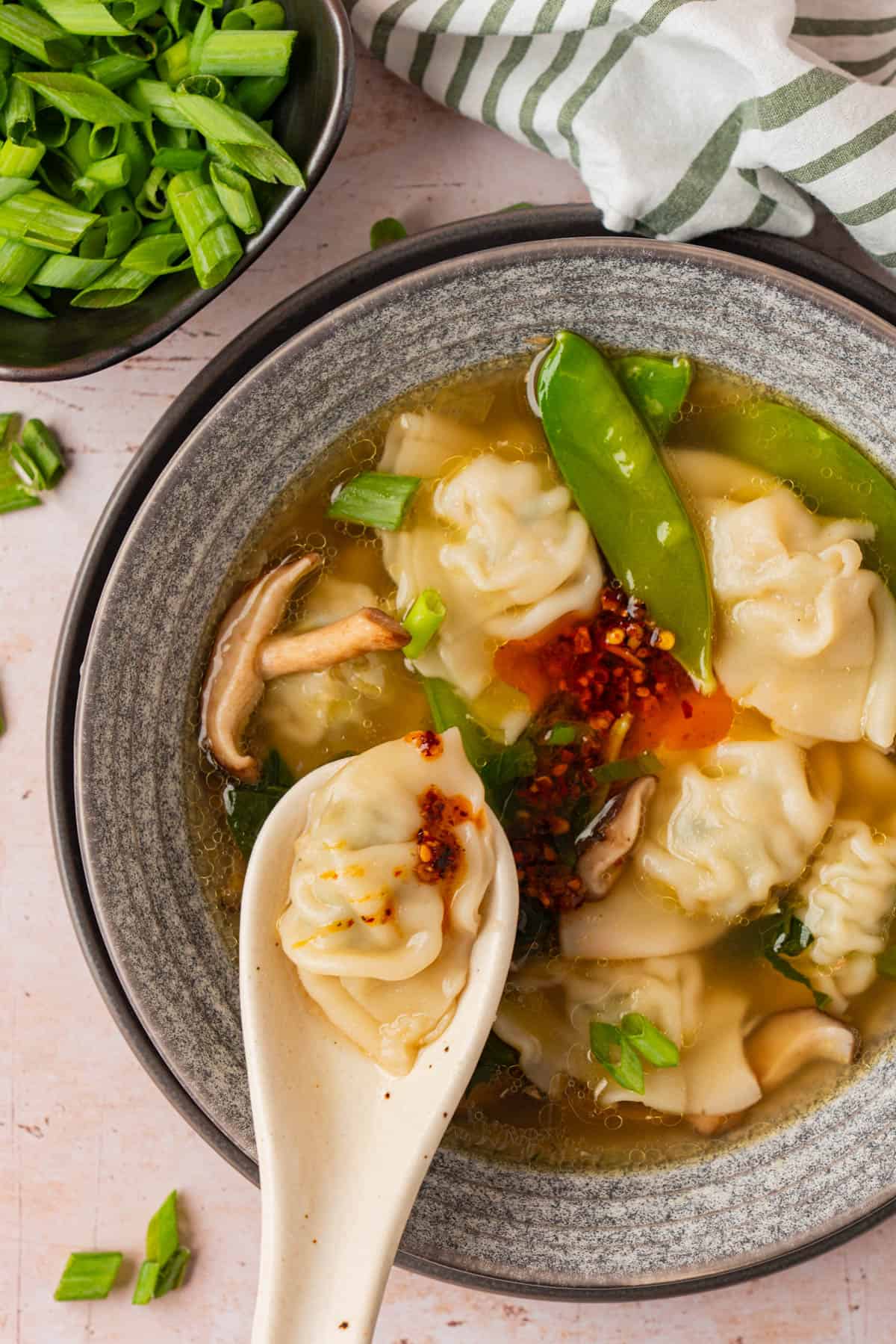 Overhead view of a bowl of potsticker soup topped with chili crisp, with a spoon lifting one potsticker out, and a bowl of chopped green onions alongside.