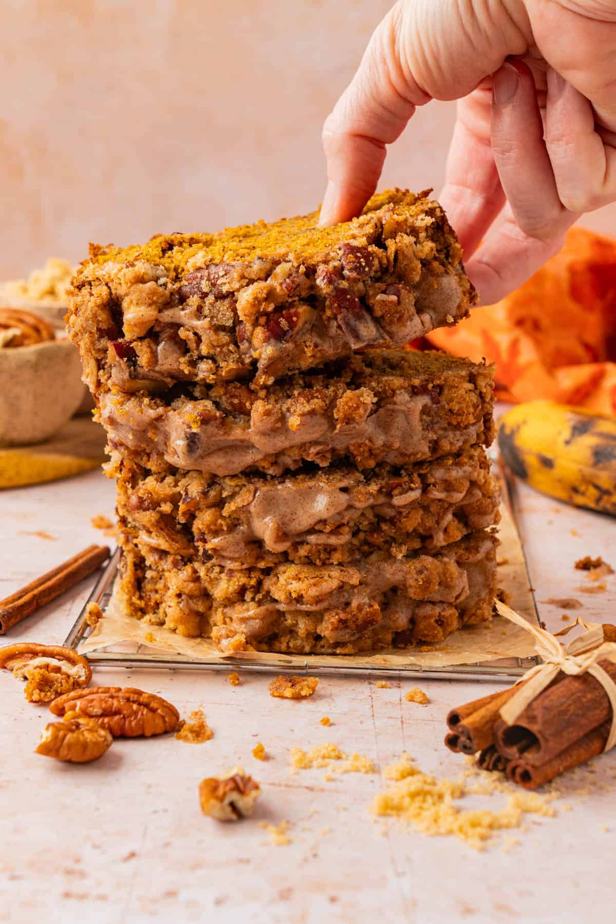 Hand lifting a slice of pumpkin banana bread from a stack of glazed slices with pecan streusel topping, cinnamon sticks and a banana in the background