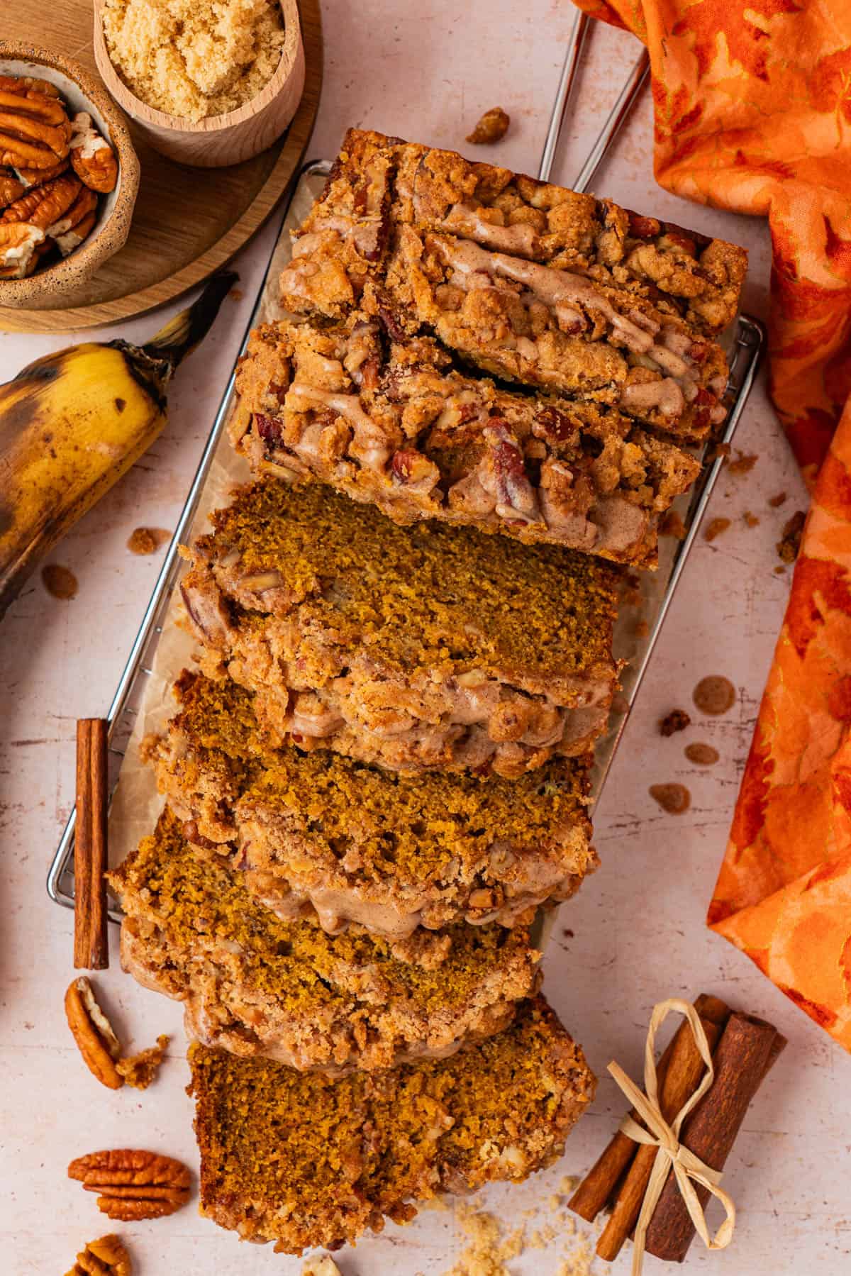 Overhead shot of sliced pumpkin banana bread with pecan streusel topping on a wire rack, surrounded by ripe bananas, pecans, brown sugar, and cinnamon sticks