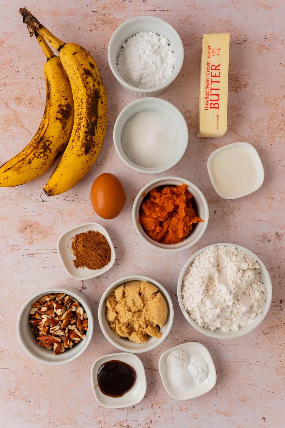 Ingredients for pumpkin banana bread laid out including ripe bananas, pumpkin puree, flour, brown sugar, powdered sugar, egg, butter, chopped pecans, cinnamon, and vanilla