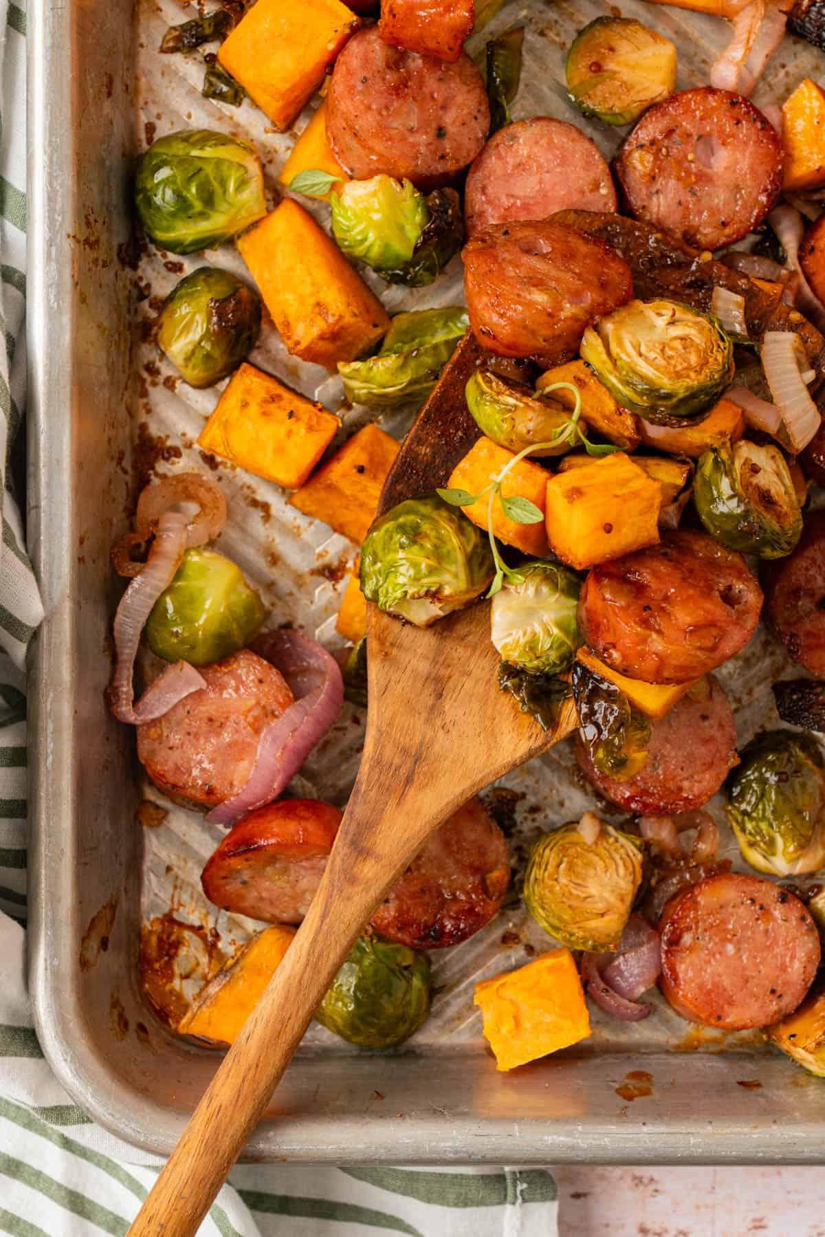 Wooden spoon scooping glazed chicken sausage, sweet potato cubes, and roasted Brussels sprouts from a sheet pan.