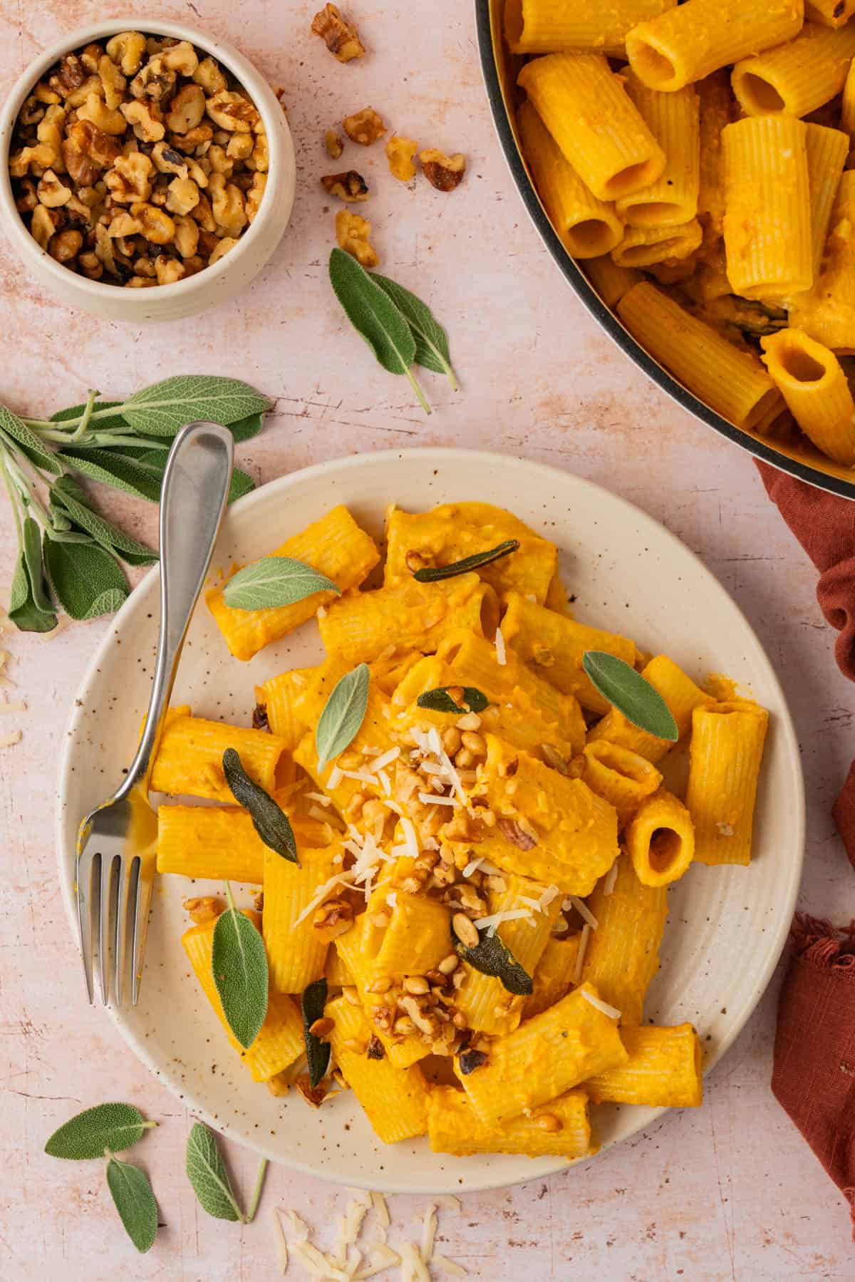 A plate of easy pumpkin pasta with walnuts, sage, and grated cheese next to a skillet of pasta with pumpkin sauce.