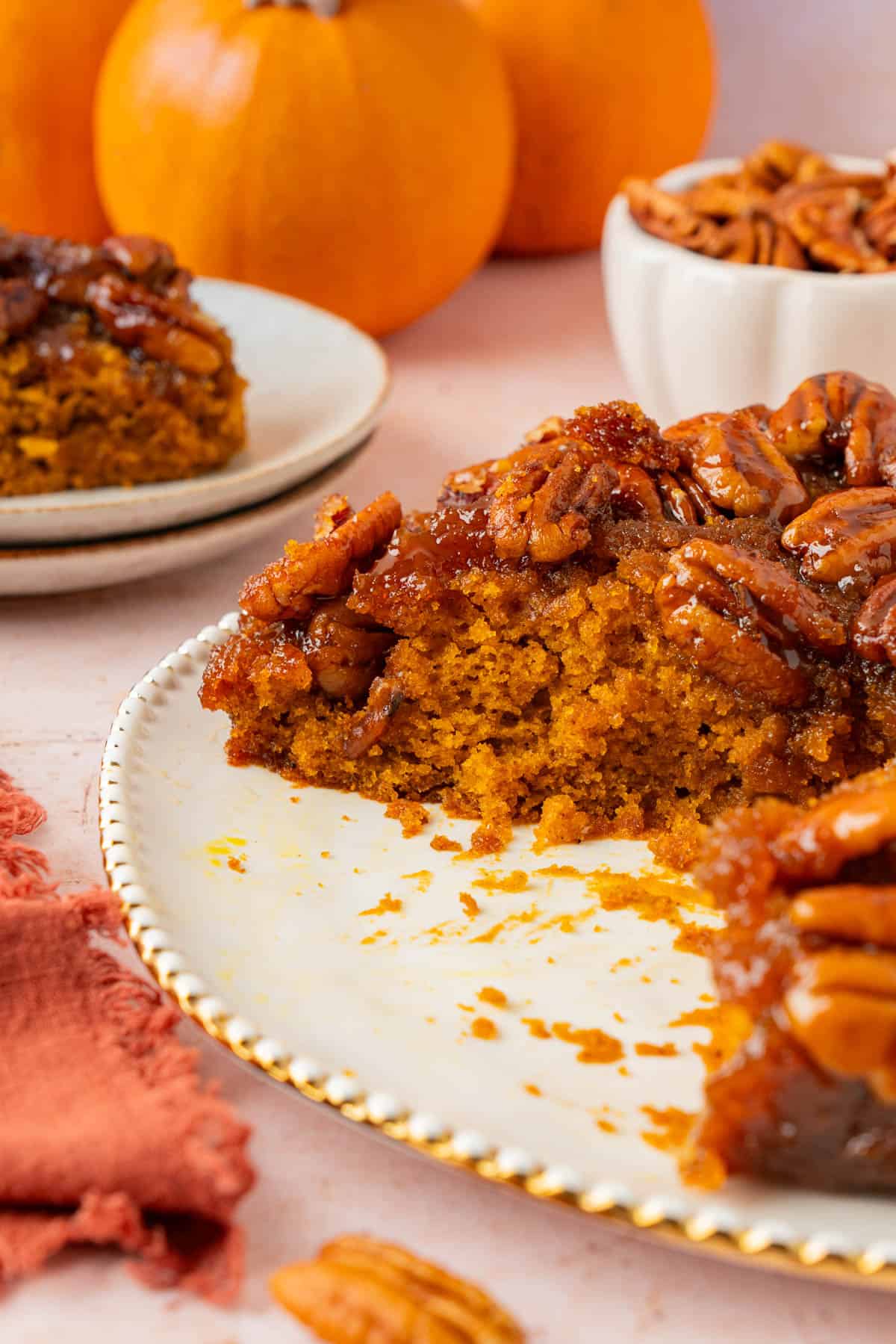 pumpkin upside down cake with a missing sliced. the missing sliced in the background, a small bowl of pecan, and a pumpkin.