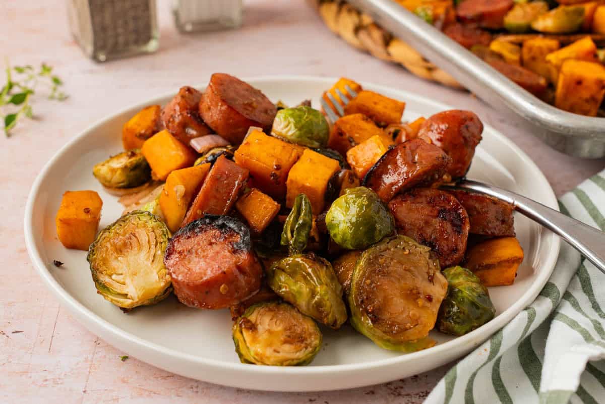 Chicken sausage and veggies sheet pan dinner served on a white plate with roasted sweet potatoes and Brussels sprouts.