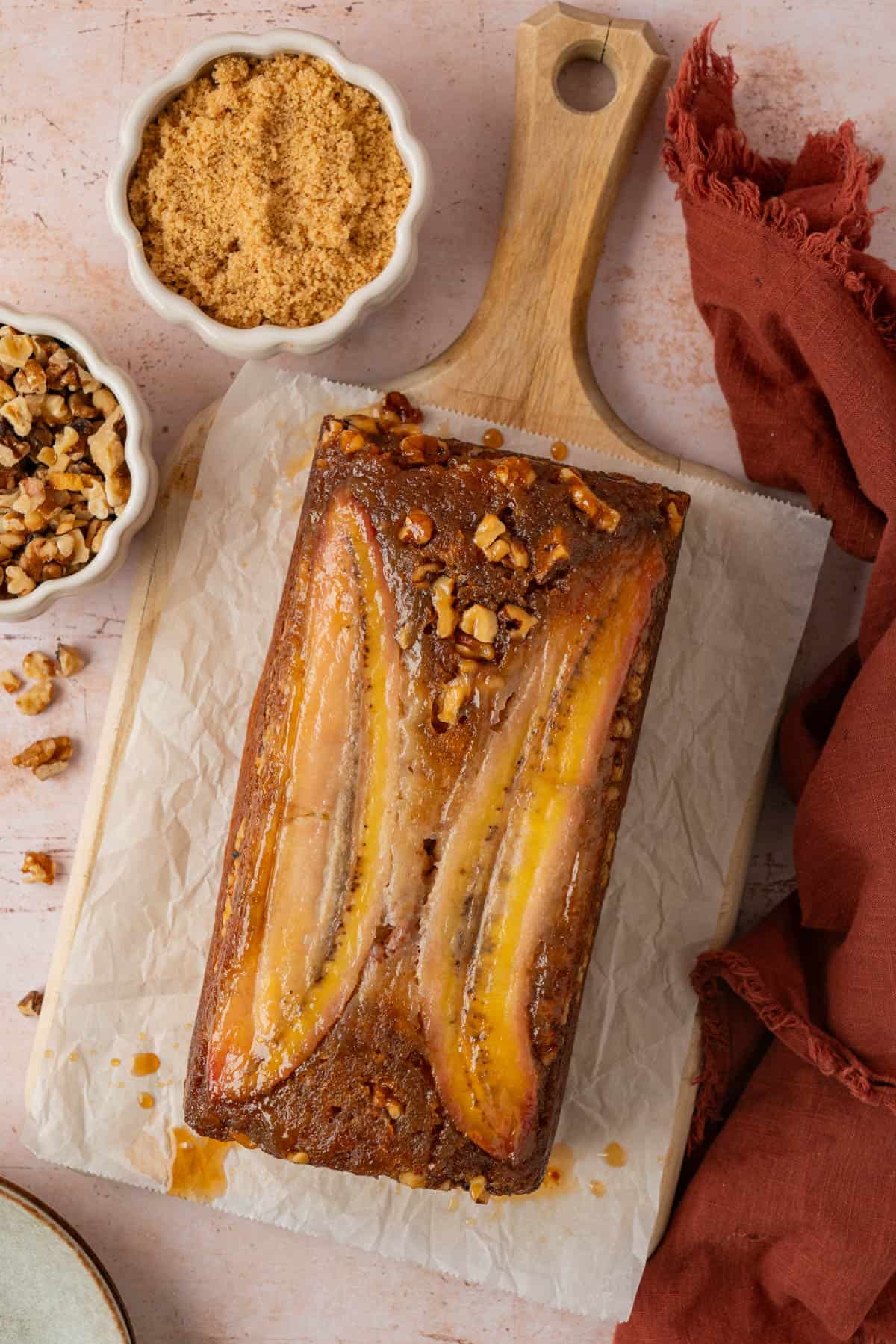 Upside down banana bread recipe topped with caramelized banana halves and walnuts on a wooden board