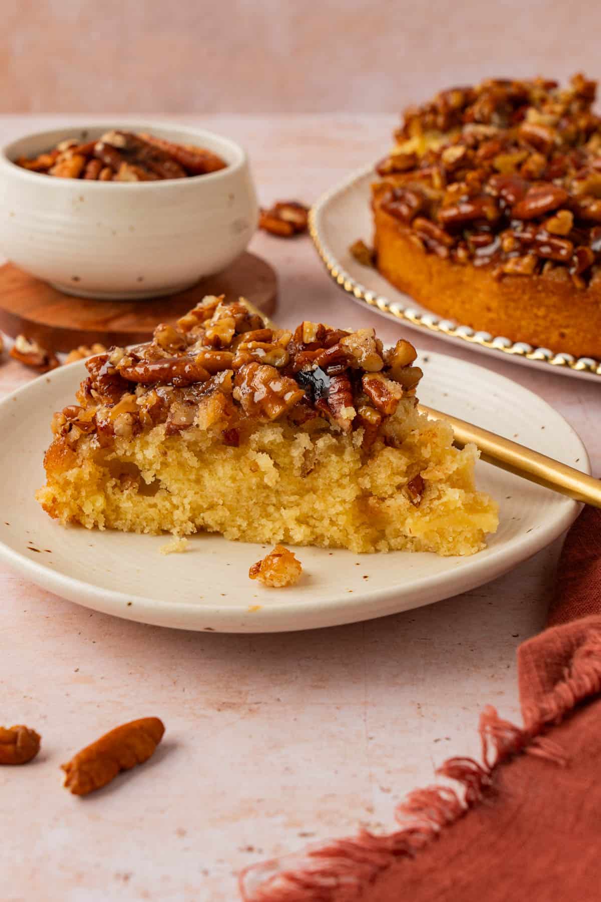 A slice of the easiest pecan upside down cake on a speckled plate with a gold fork, with the whole cake in the background.