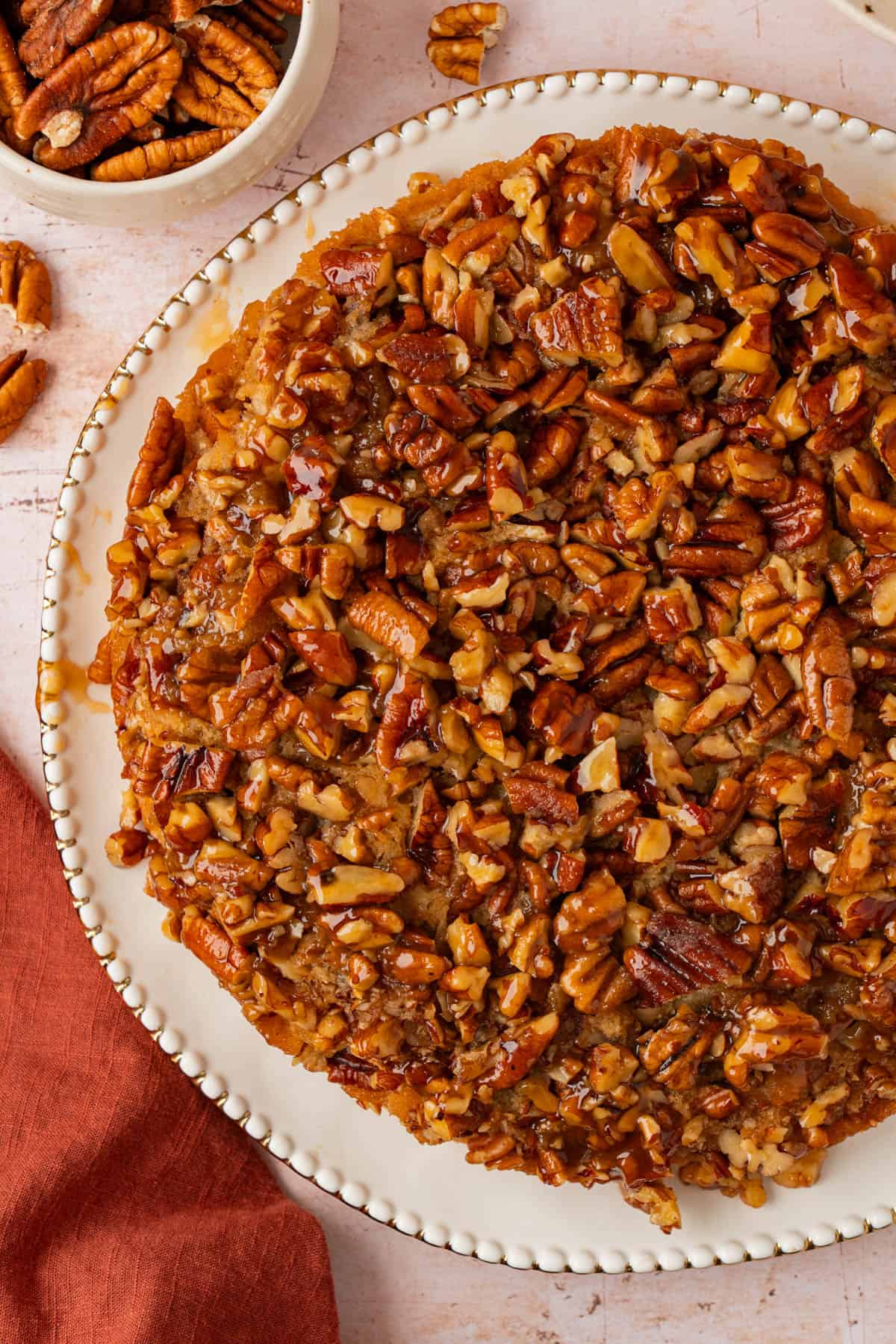 Upside down pecan cake with a gooey caramel pecan topping on a white plate with a gold beaded rim, beside a bowl of pecans.